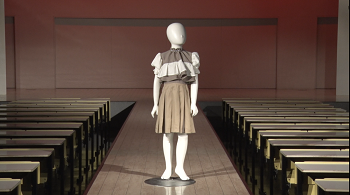 写真:マネキンが、茶色と白のチェックに、大きな白いフリルをあしらい袖の膨らんだブラウスを着て、茶色のスカートをはいている。