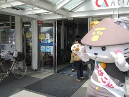 写真:笠をかぶった着ぐるみのネコが、建物の前で両手を挙げて手を振っている